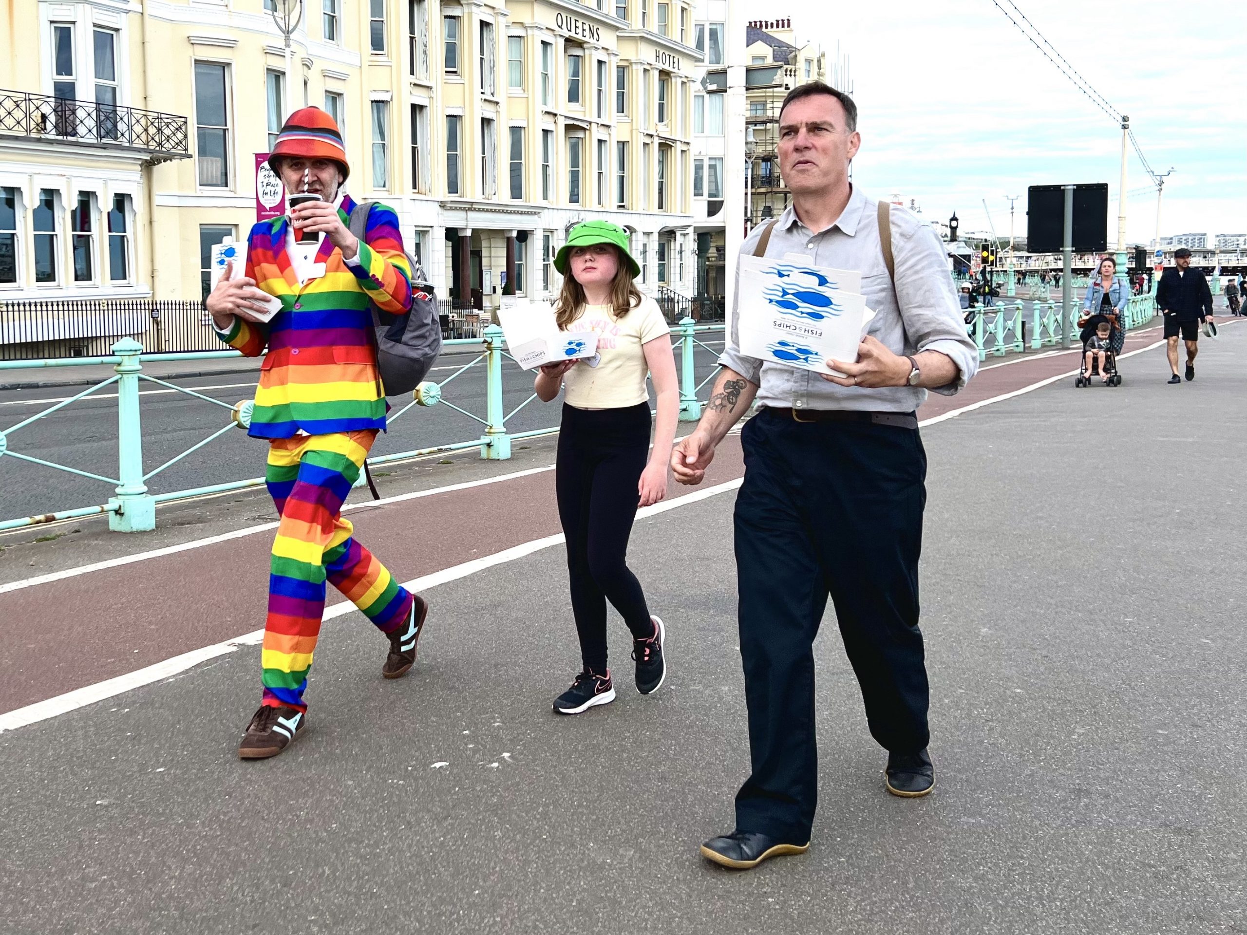 Photo of man in rainbow suit with child and man with chips walking along Brighton seafront