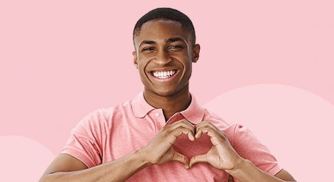 A smiling man wears a pink polo shirt. His hands are shaped into a heart.