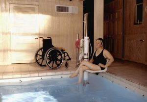 A woman is sitting on the edge of a swimming pool next to a hoist. A wheelchair is behind her.