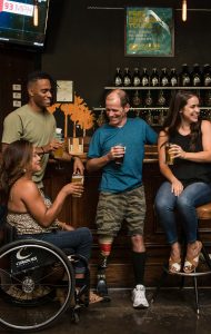 A group of 4 happy looking people are stood at a bar with drinks in their hands. One of the men has a limb difference and one of the women is seated in a wheelchair.