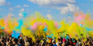 A large crowd are seen from a distance under a summer sky. They are throwing brightly coloured dust into the air
