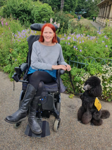 A smiling woman with bobbed red hair sits in a powered wheelchair. Next to her is a large black poodle dog with an assistance dog jacket on. They are sat in a pretty garden.