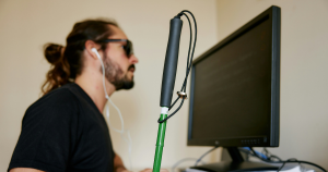a close up shot of a visually impaired persons guide cane with the user sat behind it on a computer
