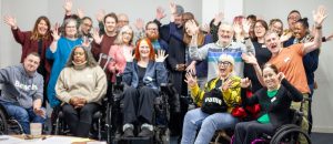 A group of around 30 people are in a large room. Some are seated in wheelchairs, some are standing. Everyone is smiling and waving and looks happy. They look like they're having a fun day.
