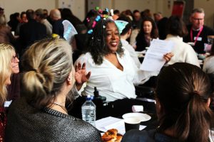 30 or 40 people are sat close together grouped round tables in a large room. We can only see the back of some people, but the faces we do see look happy and engaged. In the centre of the photo a smiling woman is seated, wearing fairy wings and holding a piece of paper. Those nearest are turned towards her and smiling.