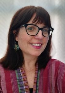 A smiling woman is half turned towards the camera. She has dark framed glasses and dark hair.