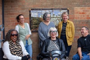 A group of 6 people are gathered in a group facing the camera, they are outside with a red brick wall behind them. 3 are seated and 3 are standing. They are all looking at each other and laughing, as if someone has just made a very good joke. They seem very happy and relaxed.