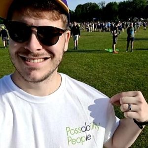 A smiling man wearing sunglasses squints at the sun. He is wearing a Possability People tshirt. Behind him we see a large park with lots of people wearing running clothes.