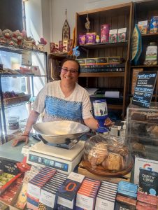 A smiling woman holds up a Possability People collection tin. Behind her we we see the interior of a shop. There are all sorts of tasty looking treats and baked goods stacked up behind her. 