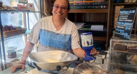 A smiling woman holds up a Possability People collection tin. Behind her we we see the interior of a shop. There are all sorts of tasty looking treats and baked goods stacked up behind her.